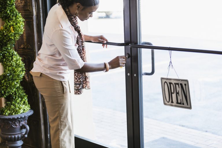 An African-American woman opening or closing a store, key in the lock in the door at the entrance. The door is glass, letting the sunlight through.
